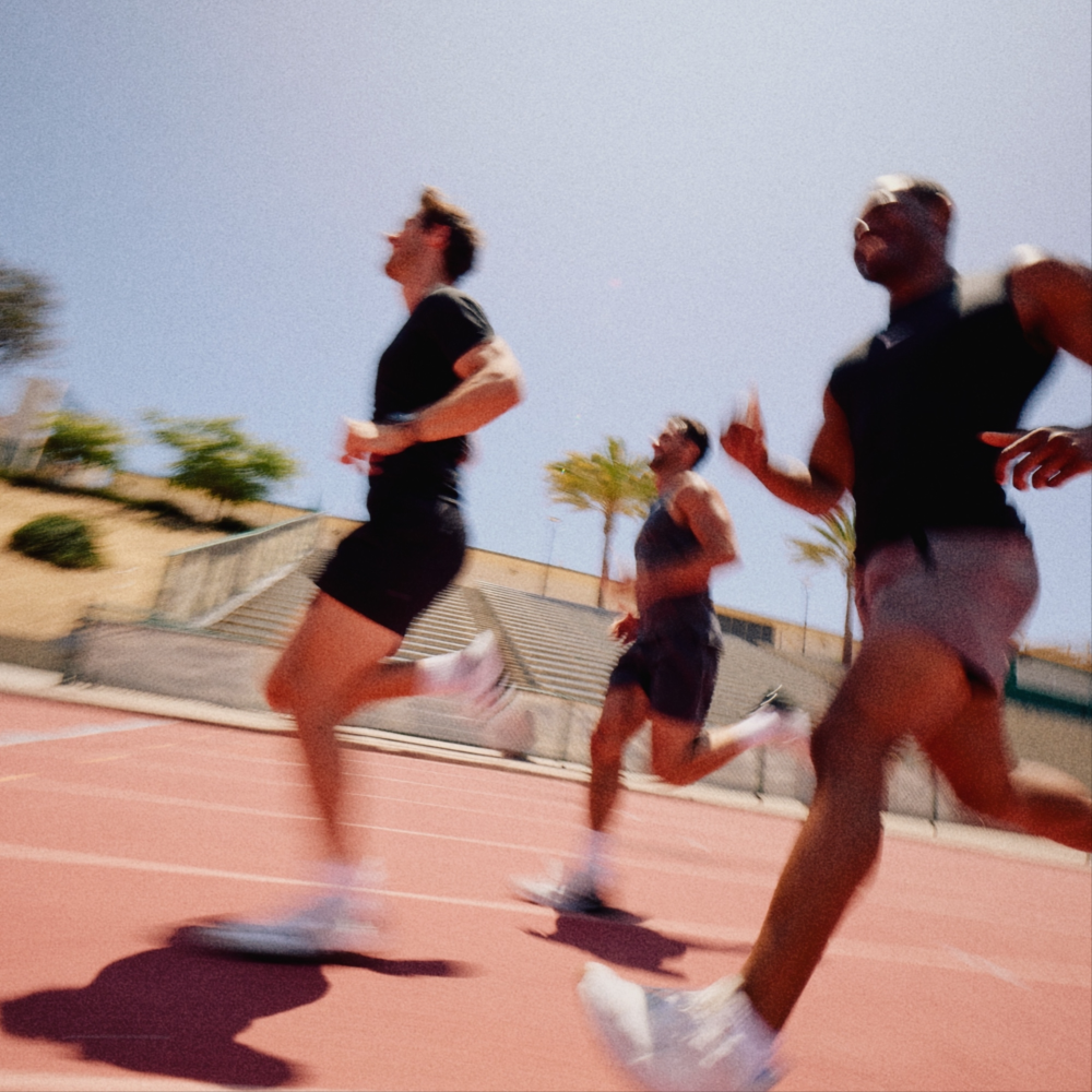 Three male athletes running on an outdoor athletic track under a clear blue sky, wearing black athletic shirts and shorts with white sneakers.