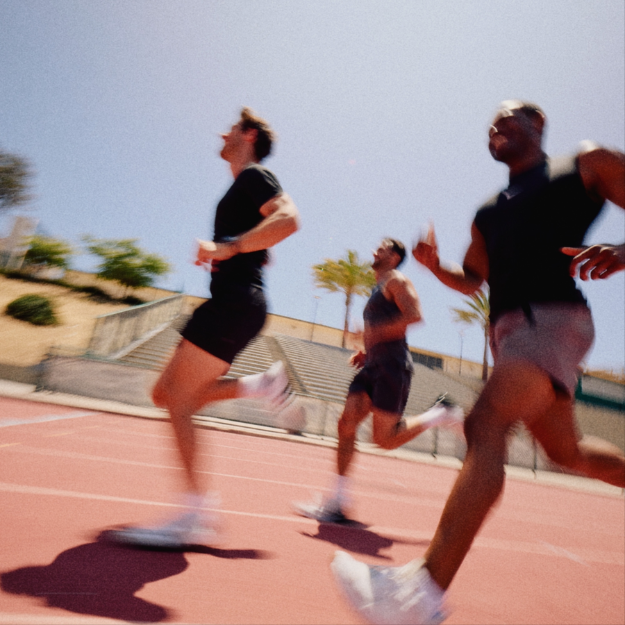 Three male athletes running on an outdoor athletic track under a clear blue sky, wearing black athletic shirts and shorts with white sneakers.