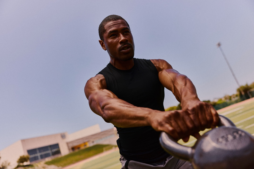 A muscular man in a black sleeveless shirt performing a kettlebell swing outdoors on a sunny day, with a blurred sports field and building in the background.