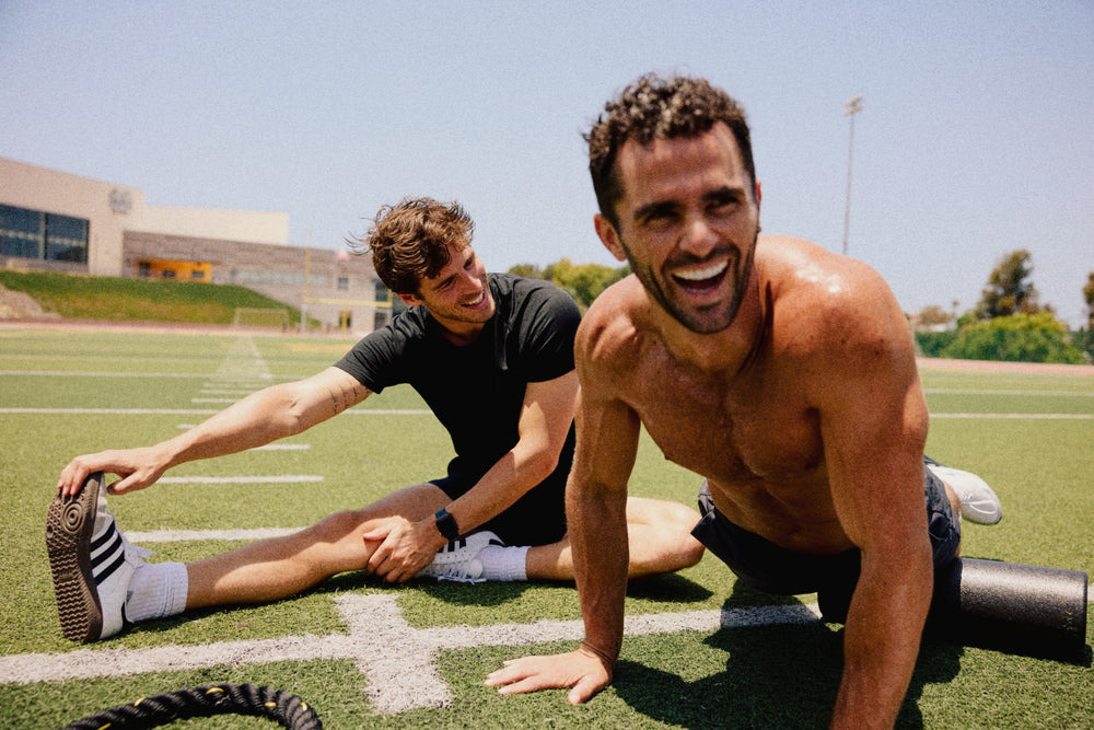 Two men stretching on a green athletic field under a clear blue sky. One shirtless man in black shorts is smiling broadly in the foreground, while another man in a black t-shirt and white sneakers stretches on the turf behind him.